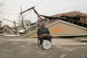 Person in front of ruined house with drum at their feet.